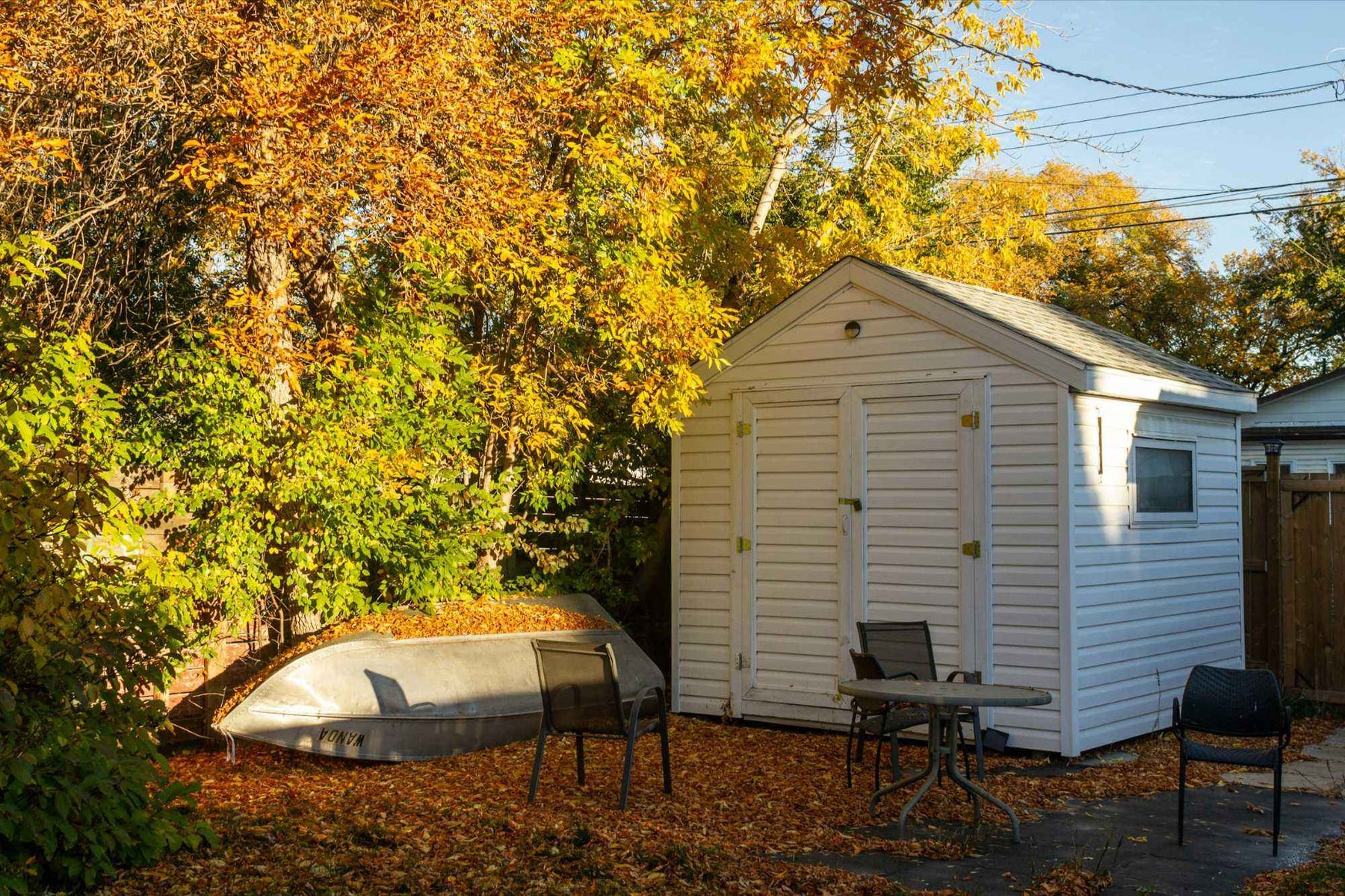 Three chairs and a round table rest on leaf-covered ground near the shed, highlighting how 7 Creative Shed Ideas can blend storage and relaxing outdoor setups