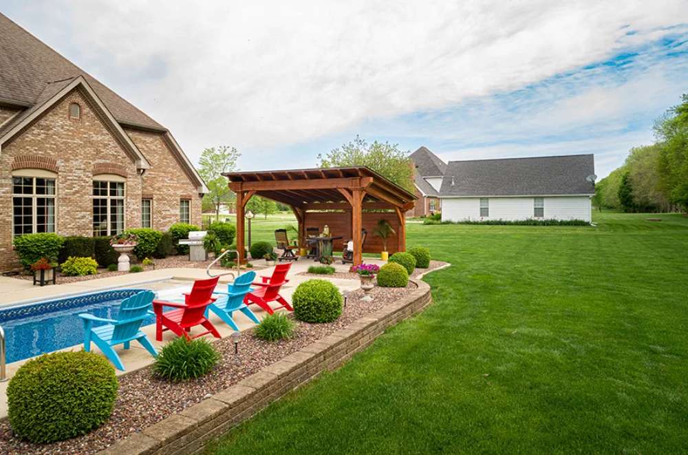 A beautiful backyard features a Brookside Timber Frame Glazier Hideaway Pavilion by the poolside, surrounded by lush greenery and colorful Adirondack chairs