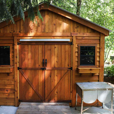 A close-up of the front of the Outdoor Living Today 12×12 Double Door Space Master Storage Shed shows rich-toned cedar wood doors, black trim hardware, and planter-style window boxes