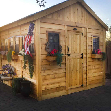 A front corner view of the Outdoor Living Today 12×16 Sunshed Garden Shed with American flag decor, window planters, and rustic charm