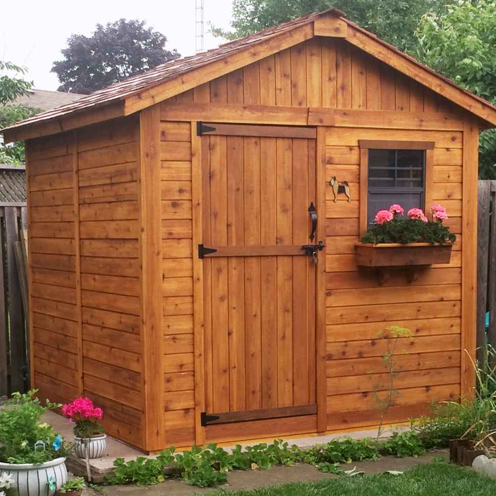A wooden shed with a dark stain, front door, and window flower box with pink flowers, standing in a garden, showing the Outdoor Living Today 8×8 Gardener Shed