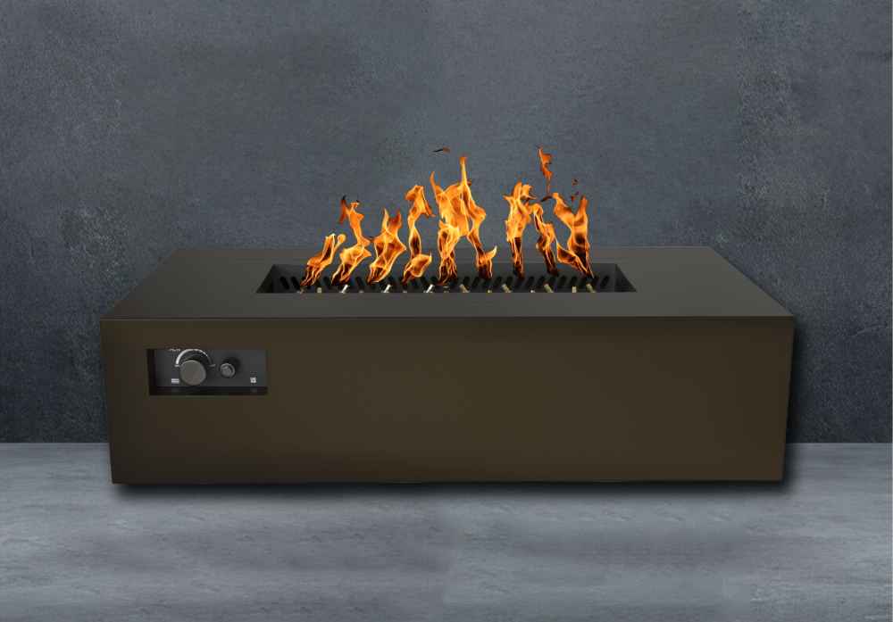 Bronze Warming Trends AON Steel Rectangular Fire Pit Table is lit with vibrant orange flames in front of a textured gray wall