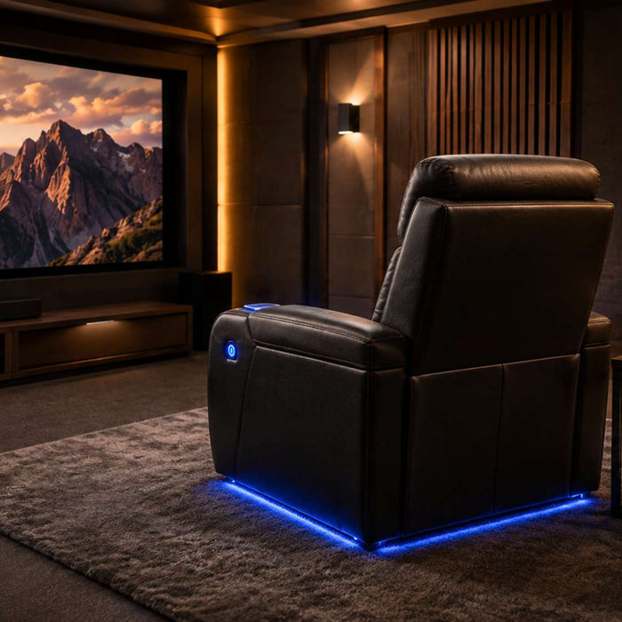 A single home theater recliner faces a large movie screen in a dark, modern media room with warm lighting, acoustic panels, and a small side table