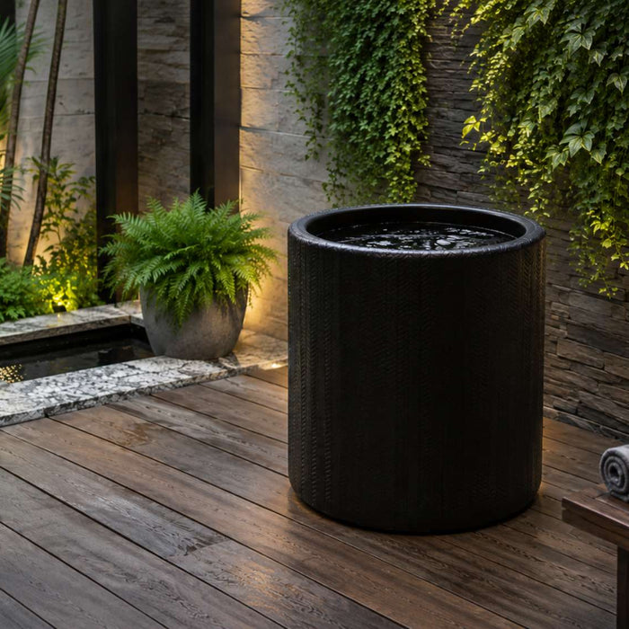 Black cold plunge barrel sits on a wooden deck in a lush outdoor wellness space with stone walls, greenery, towels, and warm accent lighting
