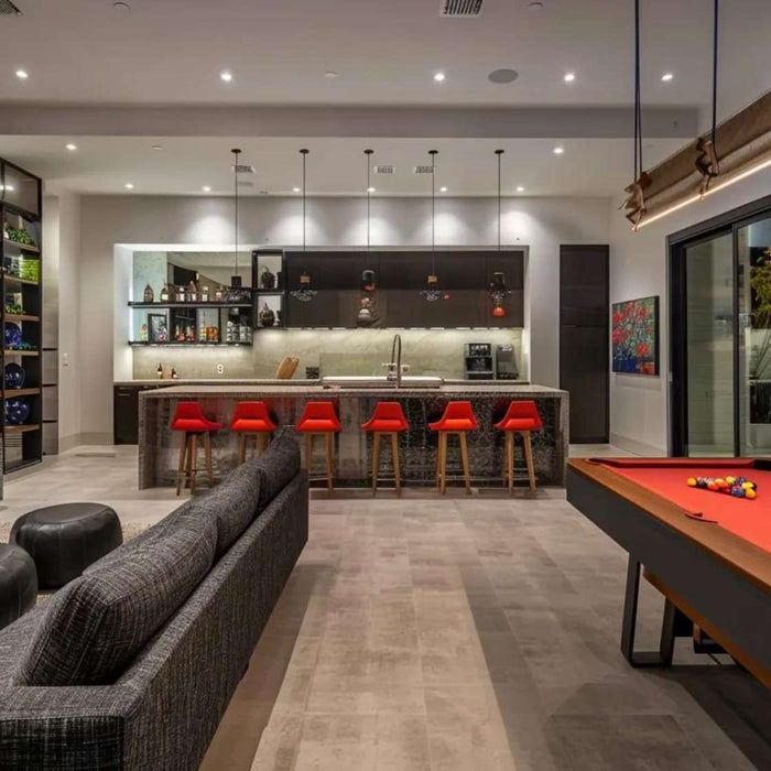 Modern game room with bar featuring red bar stools, a long marble counter, a red-felt pool table, and a cozy seating area under warm lighting