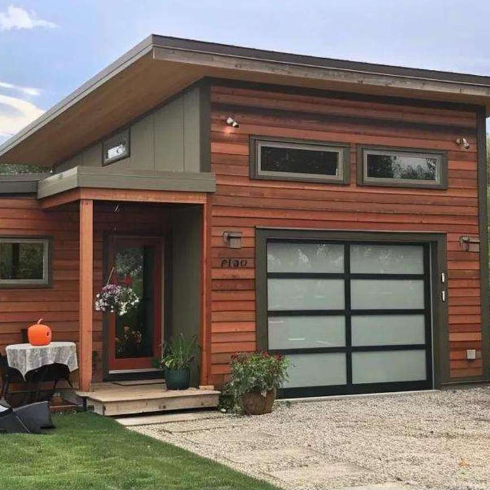 Modern wooden tiny home with garage featuring a flat roof, frosted glass garage door, front porch seating, and landscaped yard surrounded by trees