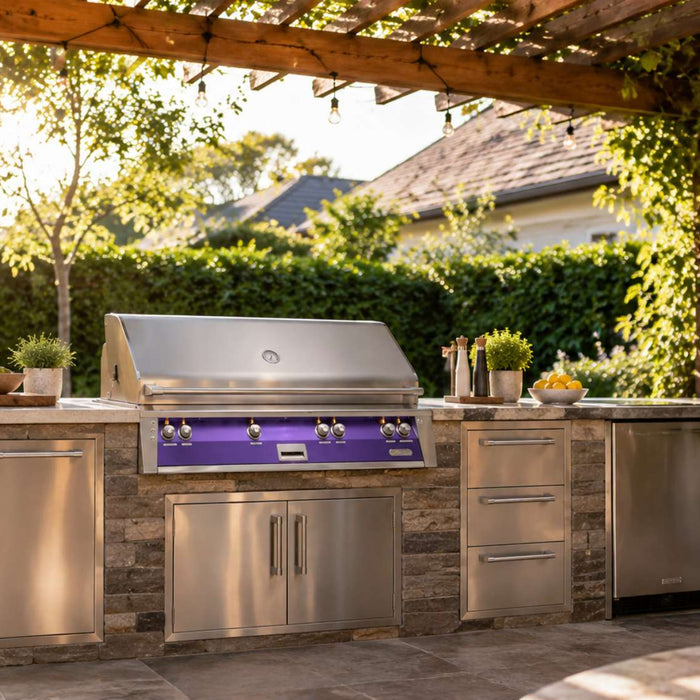 Stainless steel built in outdoor grill sits within a stone kitchen island under a wooden pergola surrounded by lush morning greenery