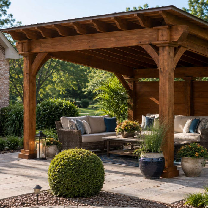 Timber frame pavilion with cushioned outdoor seating sits beside a brick home in a landscaped backyard with flowers and greenery