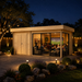 Rectangular wood cabin with glass walls at night, showing people inside watching a television on a stone patio.