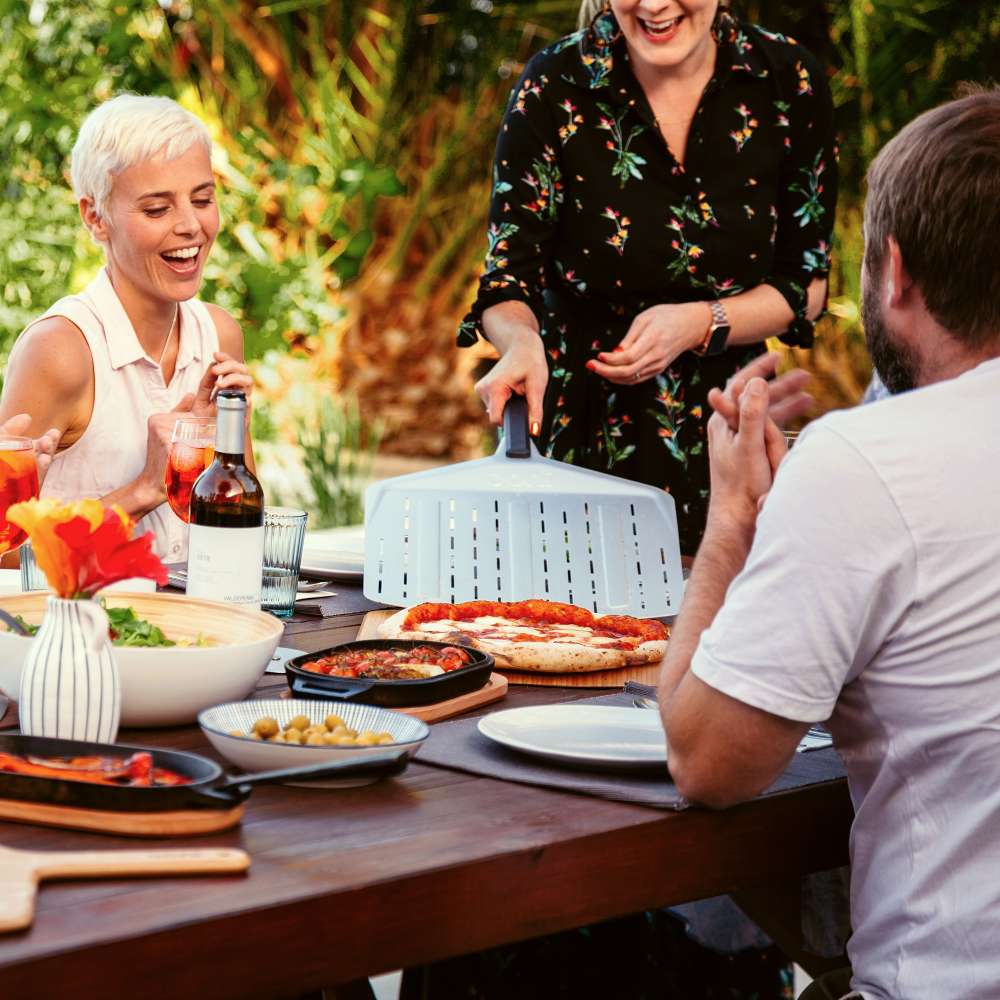 12" Ooni Perforated Pizza Peel held by a man at an outdoor dining table with friends, pizza and side dishes on the table.