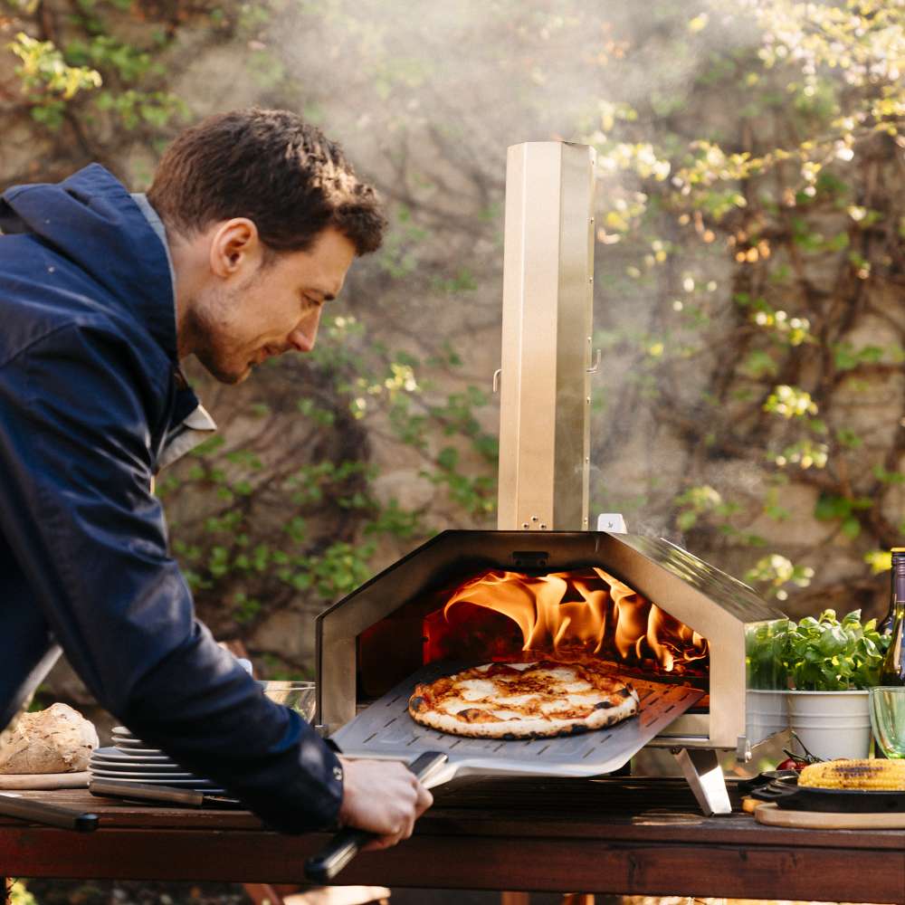 12" Ooni Perforated Pizza Peel holding a cooked pizza as a man slides it out of a lit wood fired oven with blossom behind.