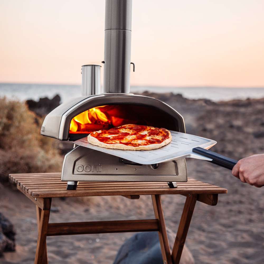 12" Ooni Perforated Pizza Peel holding a cooked pizza at the mouth of a lit oven on a beach at dusk.
