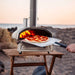 12" Ooni Perforated Pizza Peel holding a cooked pizza at the mouth of a lit oven on a beach at dusk.