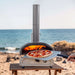 12" Ooni Perforated Pizza Peel holding a cooked pizza at the mouth of a lit oven on a wooden table with the sea behind.