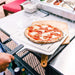 14" Ooni Perforated Pizza Peel holding an uncooked tomato and mozzarella pizza on an outdoor prep counter with a hand on the handle.
