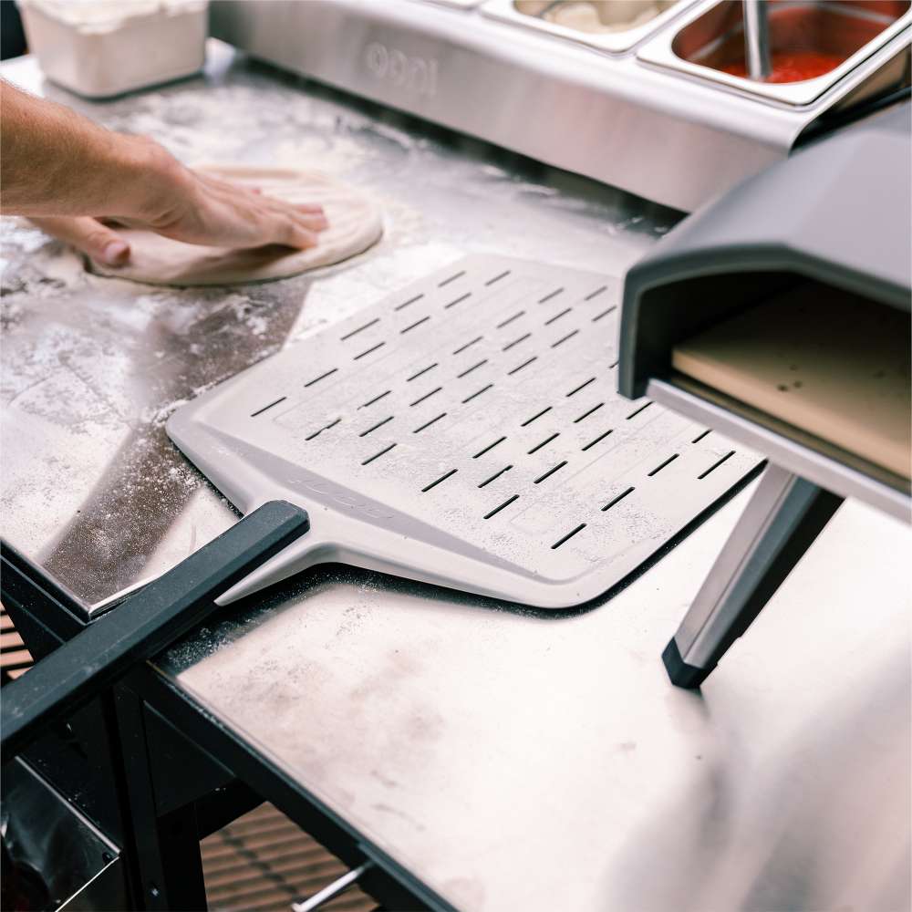 14" Ooni Perforated Pizza Peel resting on an outdoor prep station beside an oven as a hand shapes dough behind.