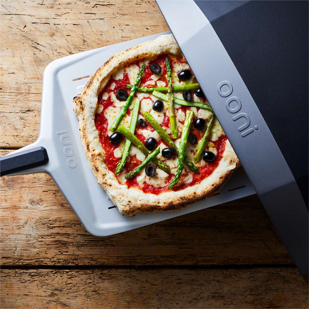 14" Ooni Perforated Pizza Peel sliding a pizza topped with asparagus and olives into an oven on a weathered wooden table.