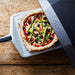 14" Ooni Perforated Pizza Peel sliding a pizza topped with asparagus and olives into an oven on a weathered wooden table.