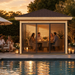 Square wood and glass pavilion with a dark roof by a pool, featuring four people dining inside at sunset.