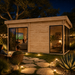 Rectangular wood studio with glass doors and windows illuminated at night in a backyard with stone path and garden lights.