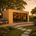 Rectangular wood studio with glass doors and a deck, illuminated at sunset with people gathered inside and outside.