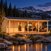 One light wood cabin with a large porch and glass doors sits by a lake at night with mountains in the background.