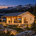 A light wood multi-room cabin with a porch at night, situated by a lake and snowy mountains.