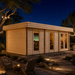 Rectangular light wood studio with flat roof and glass doors illuminated at night in a landscaped backyard.