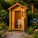 Small wood outhouse with a red gabled roof and open door revealing a white toilet, set in a garden with blue flowers.