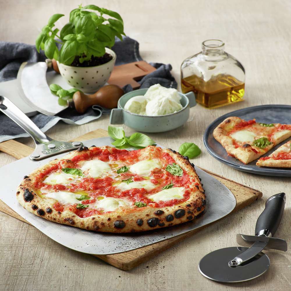 A freshly baked pizza on a peel sits beside ingredients and pizza tools made for Breville® Smart Oven® Pizzaiolo Pizza Oven