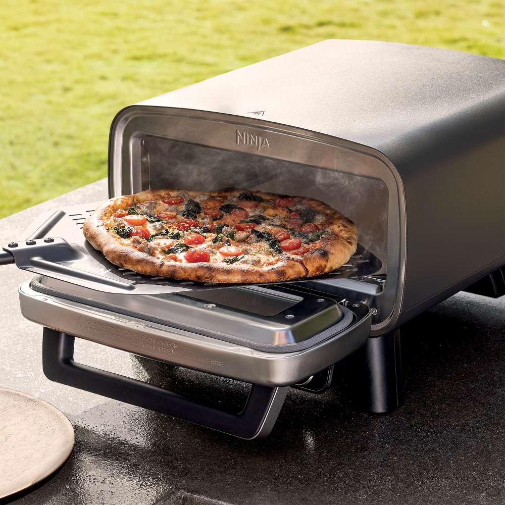 A freshly baked pizza rests on a peel in front of the open Ninja Artisan 5-in-1 Portable Electric Pizza & Outdoor Oven outdoors