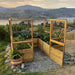 A fully planted Outdoor Living Today 8×8 Raised Garden Bed is set on a gravel base overlooking a scenic lake and mountain landscape