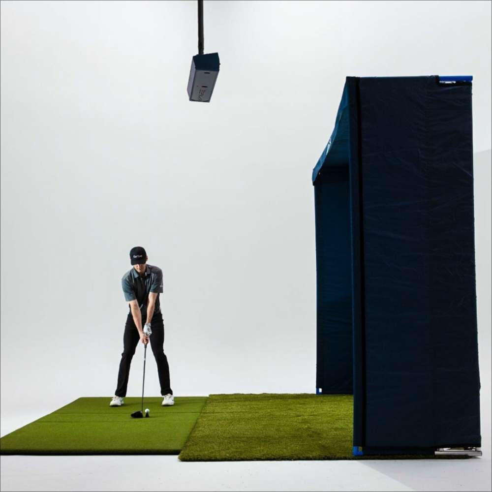 A golfer hits a ball indoors while TruGolf APOGEE Golf Launch Monitor w/ MAX Simulator tracks the swing from the ceiling above