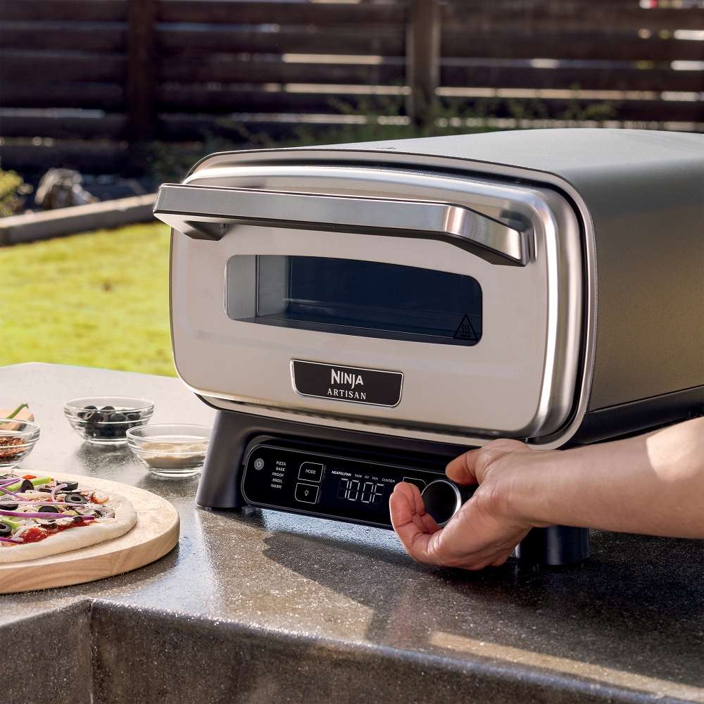 A hand adjusts the front controls of Ninja Artisan 5-in-1 Portable Electric Pizza & Outdoor Oven beside an uncooked pizza.