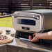 A hand adjusts the front controls of Ninja Artisan 5-in-1 Portable Electric Pizza & Outdoor Oven beside an uncooked pizza.