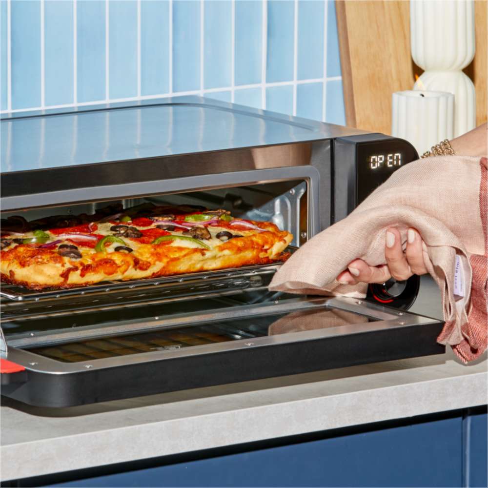 A hand removes a freshly baked pizza from the Instant Pot® InstantHeat™ Pizza Oven using a folded kitchen towel