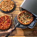 A vegetable pizza is removed with a peel from the Ninja Artisan 5-in-1 Portable Electric Pizza & Outdoor Oven beside two other pizzas