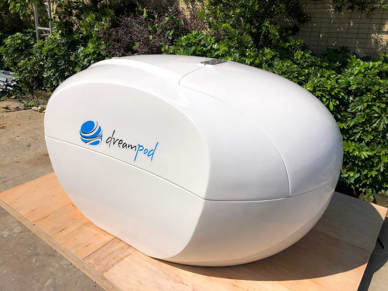 A Dreampod Home Float Plus is seen closed outdoors on a wooden platform, surrounded by lush greenery, emphasizing its smooth, capsule shape