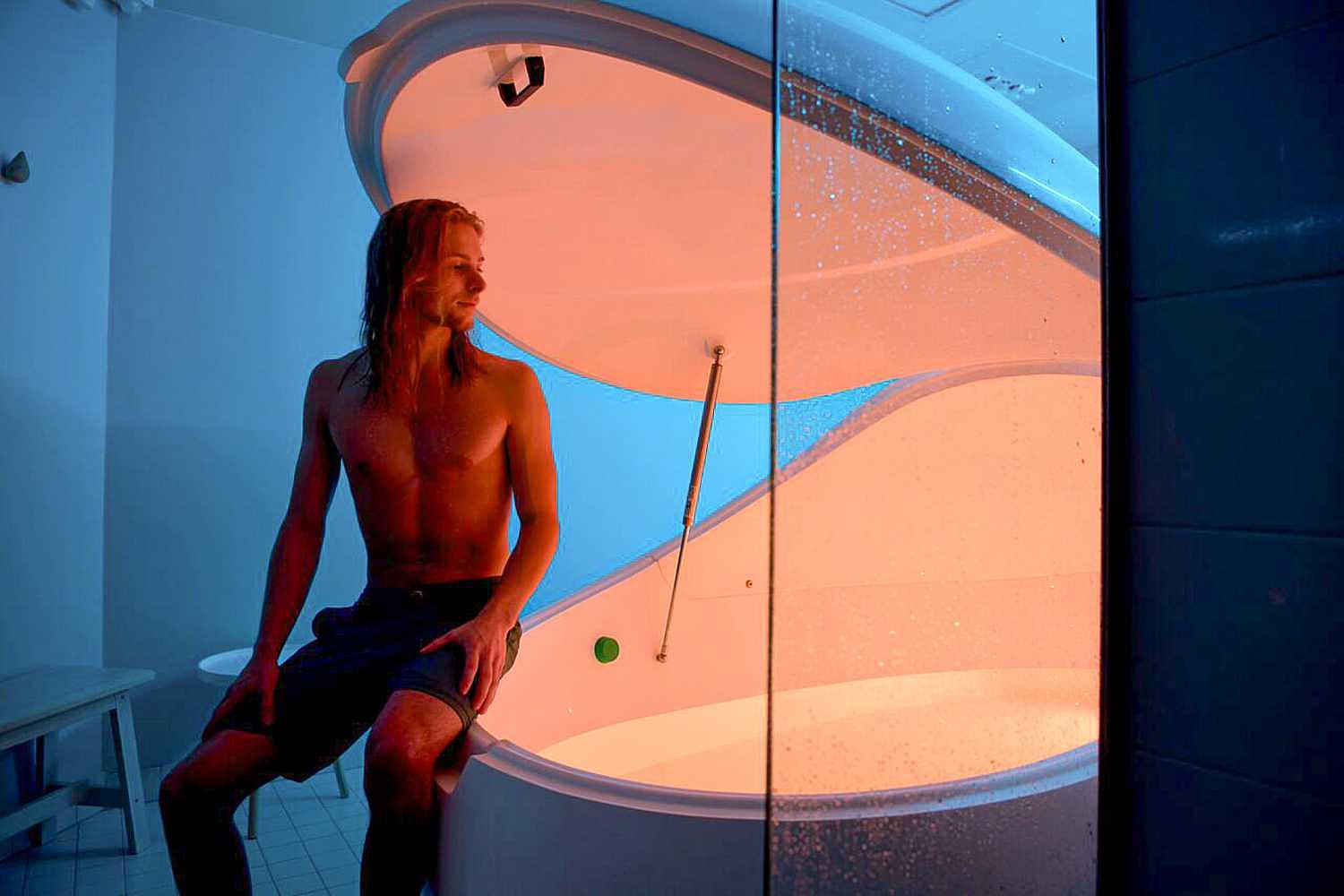 A Dreampod Vmax Float Pod with a partially open lid showing a man sitting on its edge, bathed in ambient blue and orange lighting