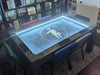 A Game Theory Tables Origins Lit setup with a black game table illuminated with a light strip and surrounded by game boxes on shelves in the background