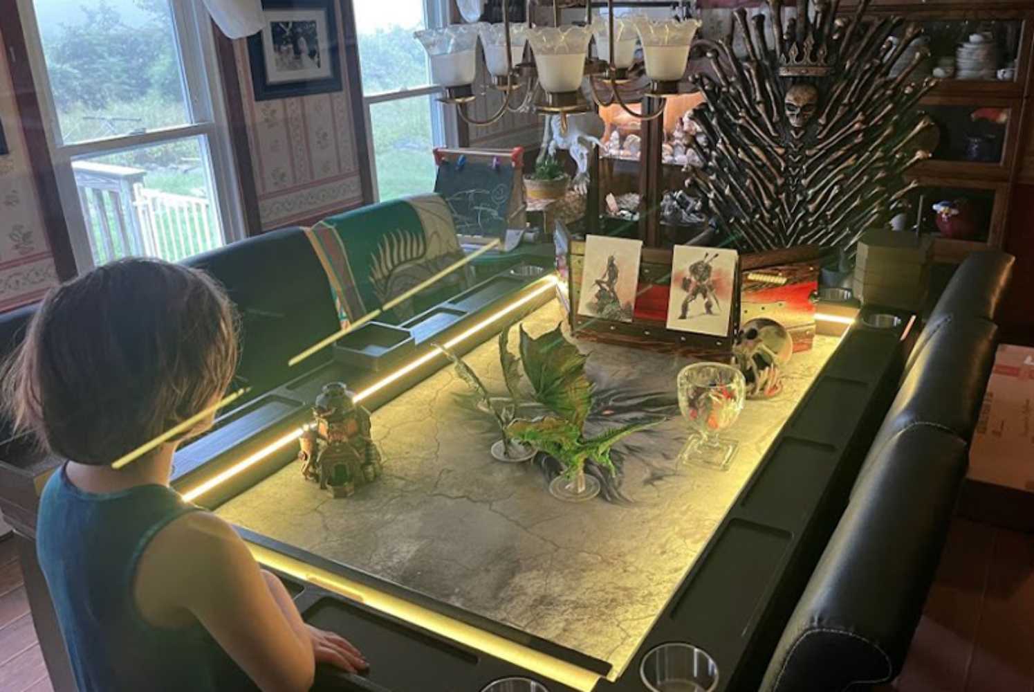 A Game Theory Tables Origins Lit table with a child observing dragon and figure miniatures, set under warm lighting