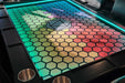A Game Theory Tables Origins Lit table with a colorful hexagon pattern in vibrant shades, bordered by bright LED lights and cup holders