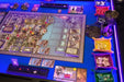 A Game Theory Tables Origins Lit table with a complex board game layout, illuminated by purple lighting and organized dice trays