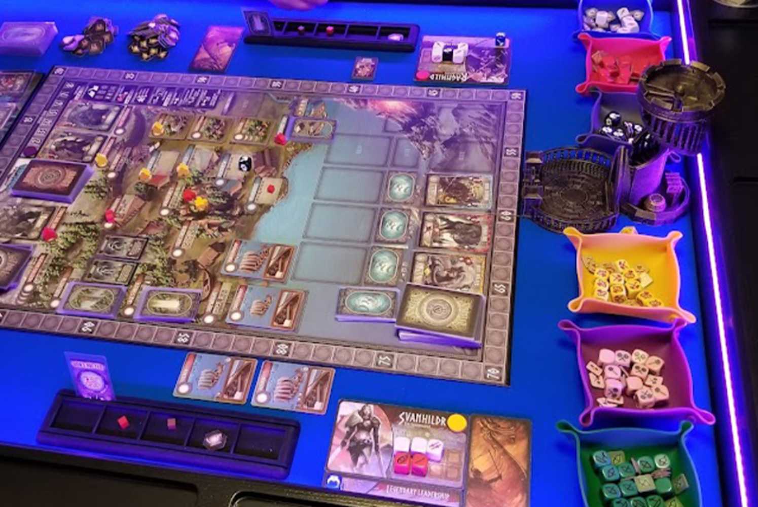 A Game Theory Tables Origins Lit table with a complex board game layout, illuminated by purple lighting and organized dice trays