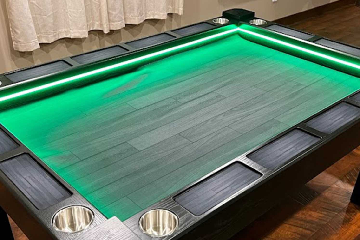 A Game Theory Tables Origins Lit table with a simple green wood-patterned surface, surrounded by cup holders and softly glowing LED lighting