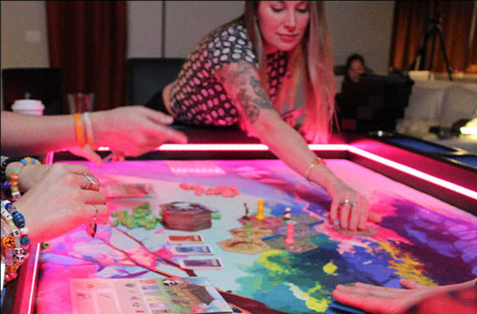 A Game Theory Tables Origins Lit table with a vibrant game scene and a woman reaching to move game pieces under pink LED lighting