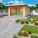 A modern wooden garden room with large sliding glass doors and a green sofa inside is shown as the GardenHouse24 USA ALU Concept 70E Garden Room