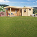 A modern, light-wood structure with large glass sliding doors opens to a furnished corner lounge, showcasing the GardenHouse24 USA ALU Concept 70N Garden Room in a blooming garden setting