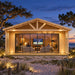 Straight-on nighttime view of the illuminated ALU Concept Arosa 44C on North Carolina’s Outer Banks, framed by sandy dunes, sea oats and coastal trees, with three adults watching a ballgame inside the glass-fronted sports room.