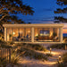 Straight-on nighttime view of the illuminated ALU Concept Lounge 44B on North Carolina’s Outer Banks, surrounded by sandy dunes, sea oats and wind-shaped coastal trees, with three adults watching a ballgame inside the glass-fronted sports room.